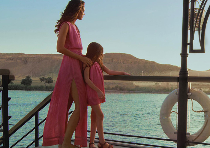 Abeer and her daughter, both in matching pink dresses, stand together, overlooking the water over a dock.