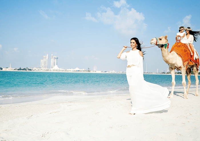 Photo, courtesy of Harper’s Bazaar Arabia, of Abeer Al Otaiba on the beach in a white dress in Abu Dhabi, pulling a camel.