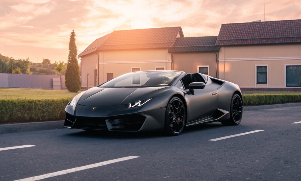 Grey Lamborghini driving on road with sunset and house in the background.